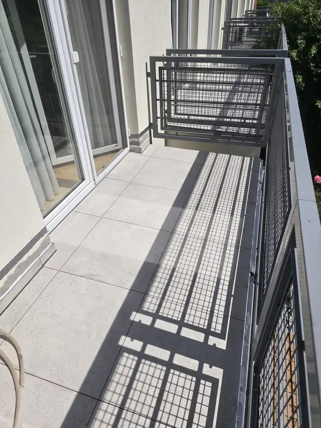 Długi balkon z jasnym gresem, balustrada z siatki rzuca wyraźny cień