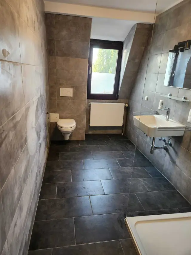 Łazienka z szarymi płytkami na ścianach, oknem i podwieszoną miską WC oraz umywalką