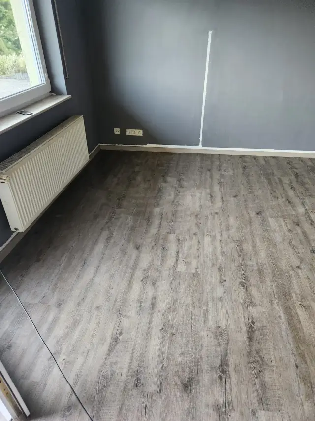 Pokój z ciemnoszarymi ścianami, podłoga z paneli imitujących drewno i grzejnik przy oknie