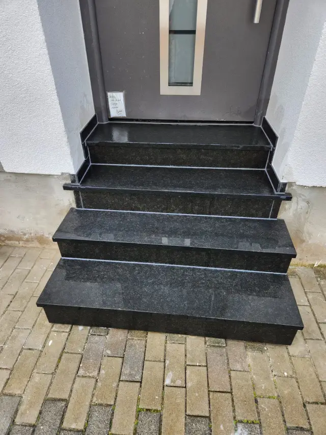 Czarne granitowe stopnie prowadzące do drzwi wejściowych, otoczone kostką brukową