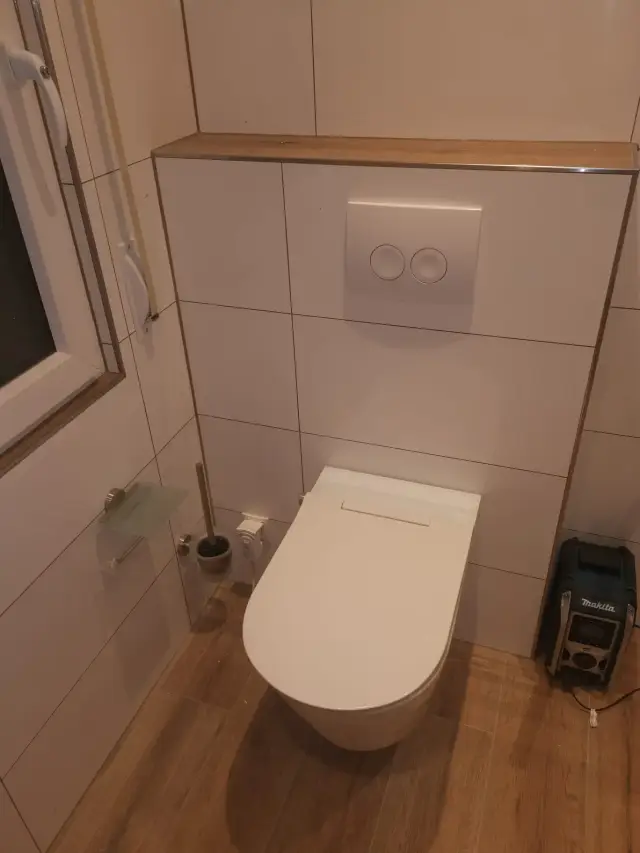 Toaleta podwieszana z przyciskiem spłukiwania w ścianie z płytek i drewnianą półką nad nią