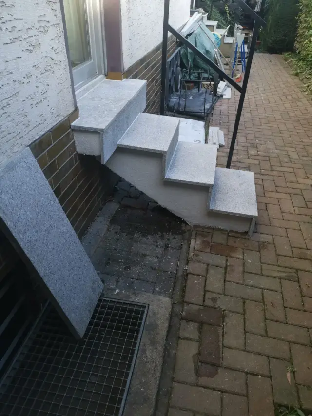 Zewnętrzne schody z jasnego granitu przy domu, obok płyta oparta o ścianę, w trakcie prac