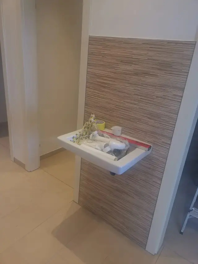 Umywalka wisząca przy ścianie z pasową płytką, na umywalce leżą materiały montażowe