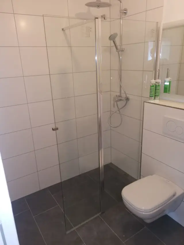 Łazienka z prysznicem za szkłem, białymi płytkami na ścianach i podwieszaną miską WC