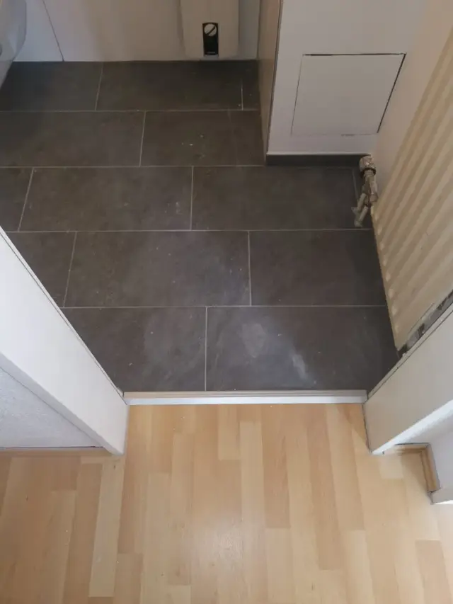 Ciemne płytki na podłodze przy progu do podłogi w imitacji drewna, grzejnik w rogu