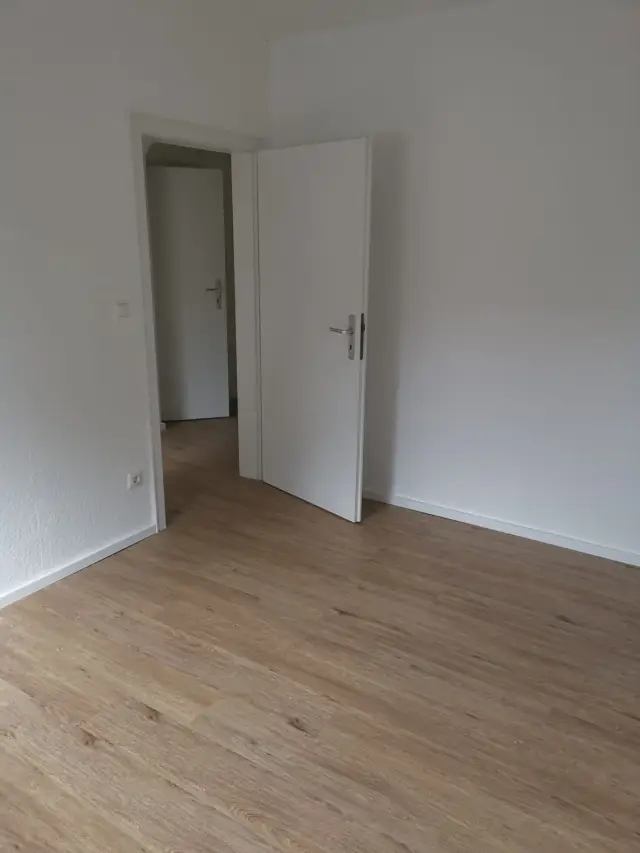 Pokój z jasną podłogą w imitacji drewna, białymi ścianami i otwartymi drzwiami