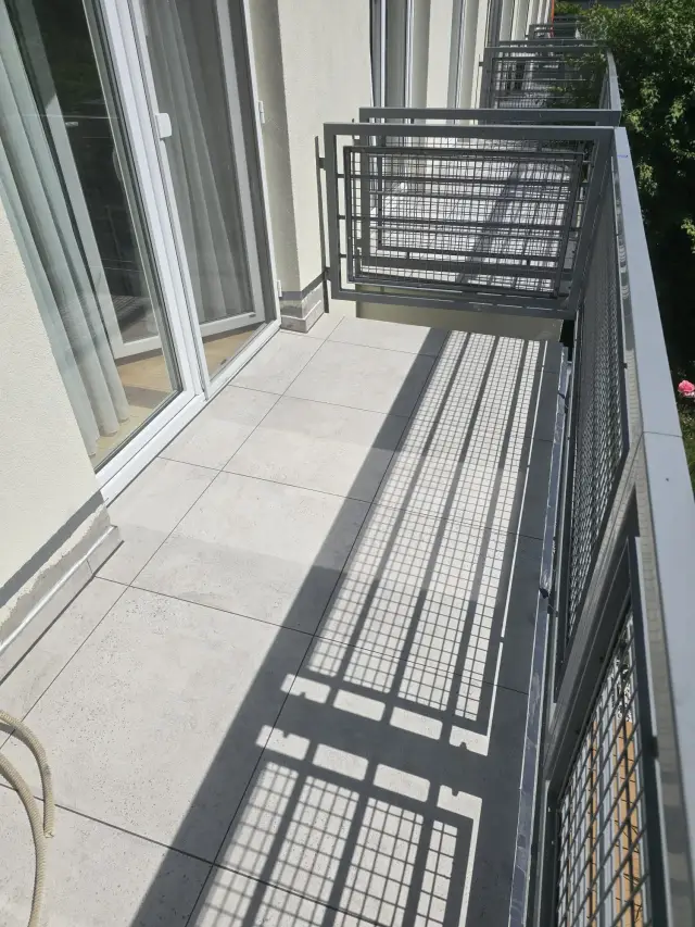 Długi balkon z jasnym gresem, balustrada z siatki rzuca wyraźny cień