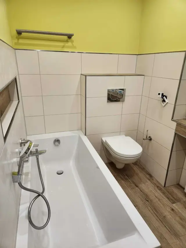 Łazienka z wanną, podwieszaną miską WC i beżowymi płytkami, żółta ściana