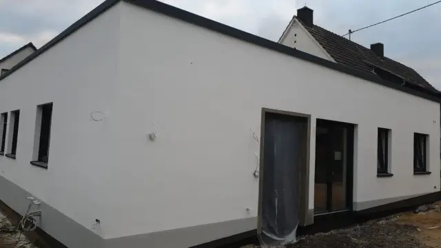 Parterowy dom z białym tynkiem i oknami w ciemnych ramach, drzwi osłonięte folią
