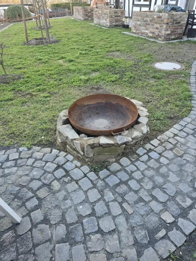 Kamienny krąg z rdzawą metalową misą na ognisko w ogrodzie, otoczony kostką brukową